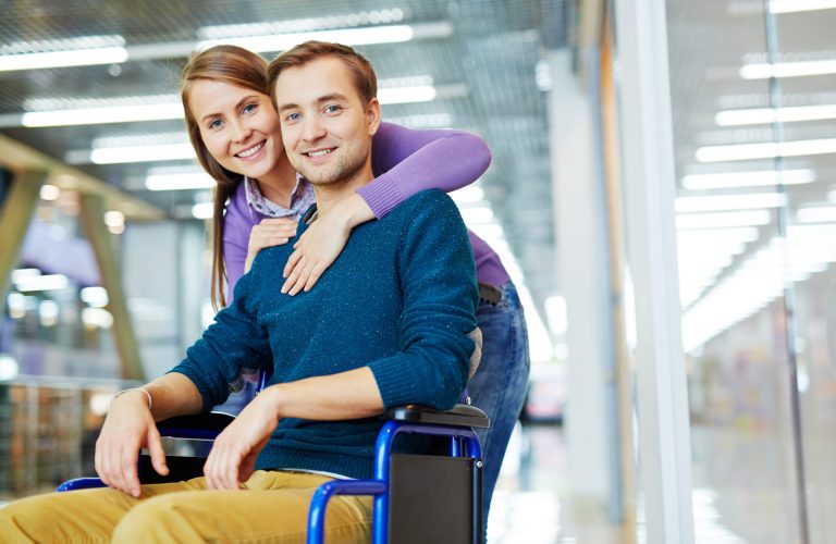 Portrait of young happy couple: pretty girl hugging her smiling handicapped boyfriend in wheelchair